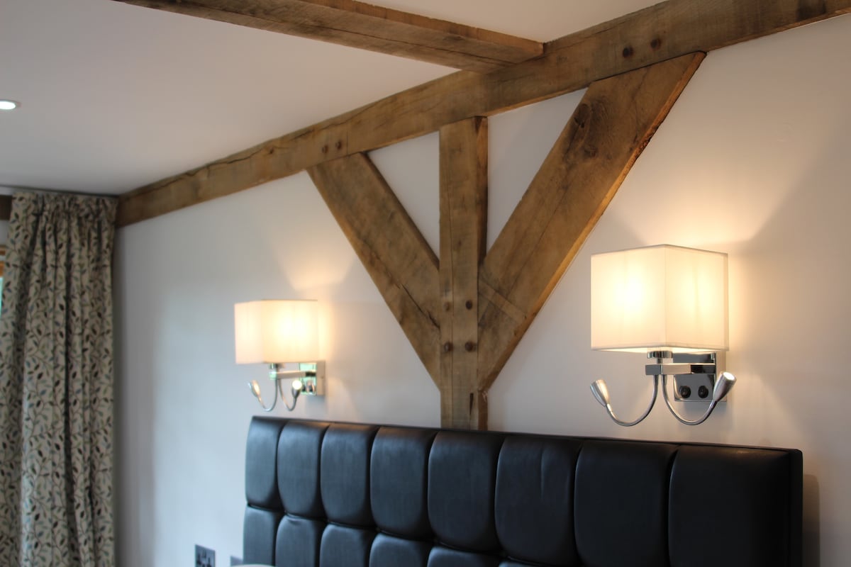 Textured wooden beams provide structure to the room, enhancing the rustic charm. Two lamps mounted on the wall offer soft lighting beside a black upholstered headboard, creating a cozy and welcoming atmosphere. Drapery featuring subtle patterns adds a touch of warmth to the space.