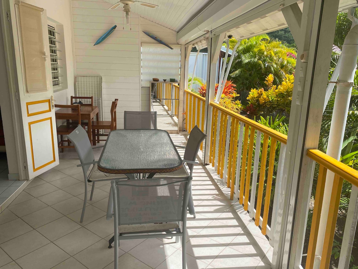 An outdoor terrace is featured, equipped with a glass-top dining table and four metal chairs. Lush greenery and colorful plants surround the area, providing a vibrant backdrop. Sunlight casts shadows on the tiled floor, enhancing the inviting space.