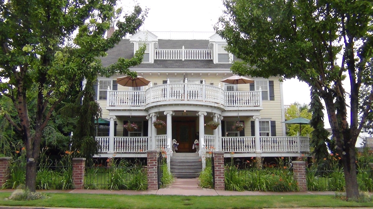 The exterior of the inn displays a charming three-story structure with a wide front porch adorned with umbrellas and hanging flower baskets. Lush greenery and landscaped flower beds frame the entrance, while tall trees provide shade, enhancing the inviting atmosphere.