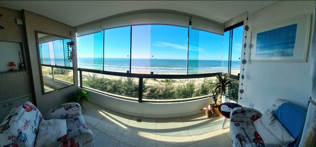 A panoramic view showcases large floor-to-ceiling windows that frame the ocean and shoreline. Two cozy chairs are positioned to enjoy the scenery, while a decorative plant adds a touch of greenery. Natural light illuminates the space, enhancing the peaceful atmosphere.