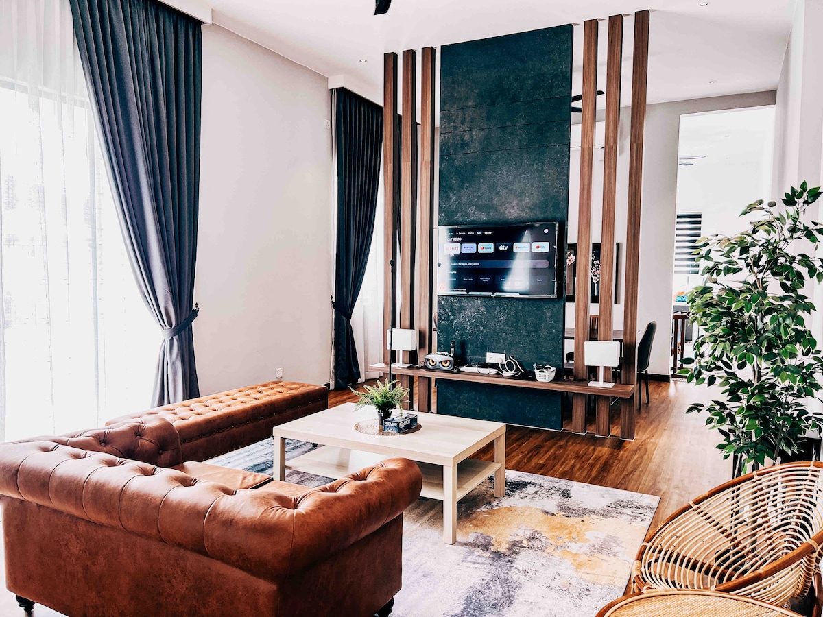 A stylish living area features a combination of leather sofas and a wooden coffee table. A large flat-screen TV is mounted on a textured wall, flanked by decorative elements. Natural light floods the space through large windows, accented by elegantly draped curtains.