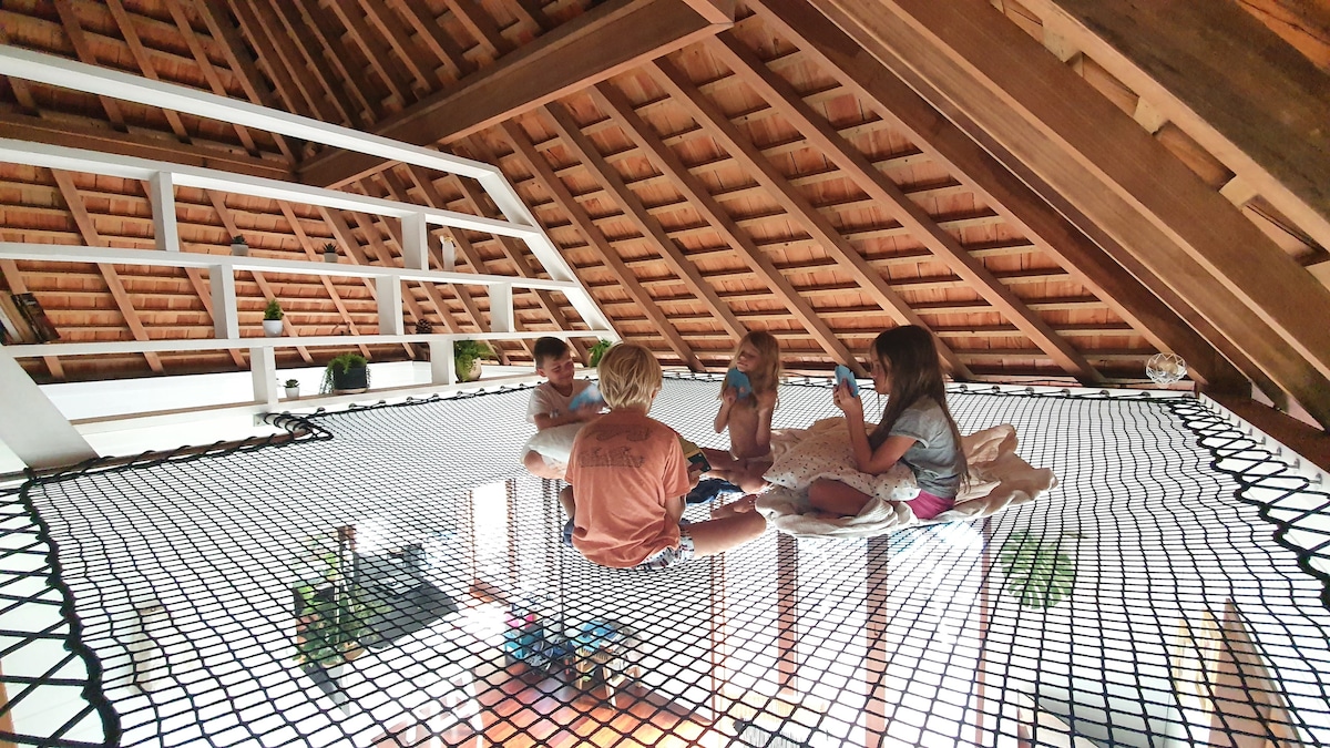 A spacious mezzanine area features a soft netting base, inviting relaxation and fun. Natural light filters through the open space, highlighting a relaxed atmosphere. Children are seated comfortably on the net, creating a lively scene of ease and enjoyment amidst the villa's wooden architecture.