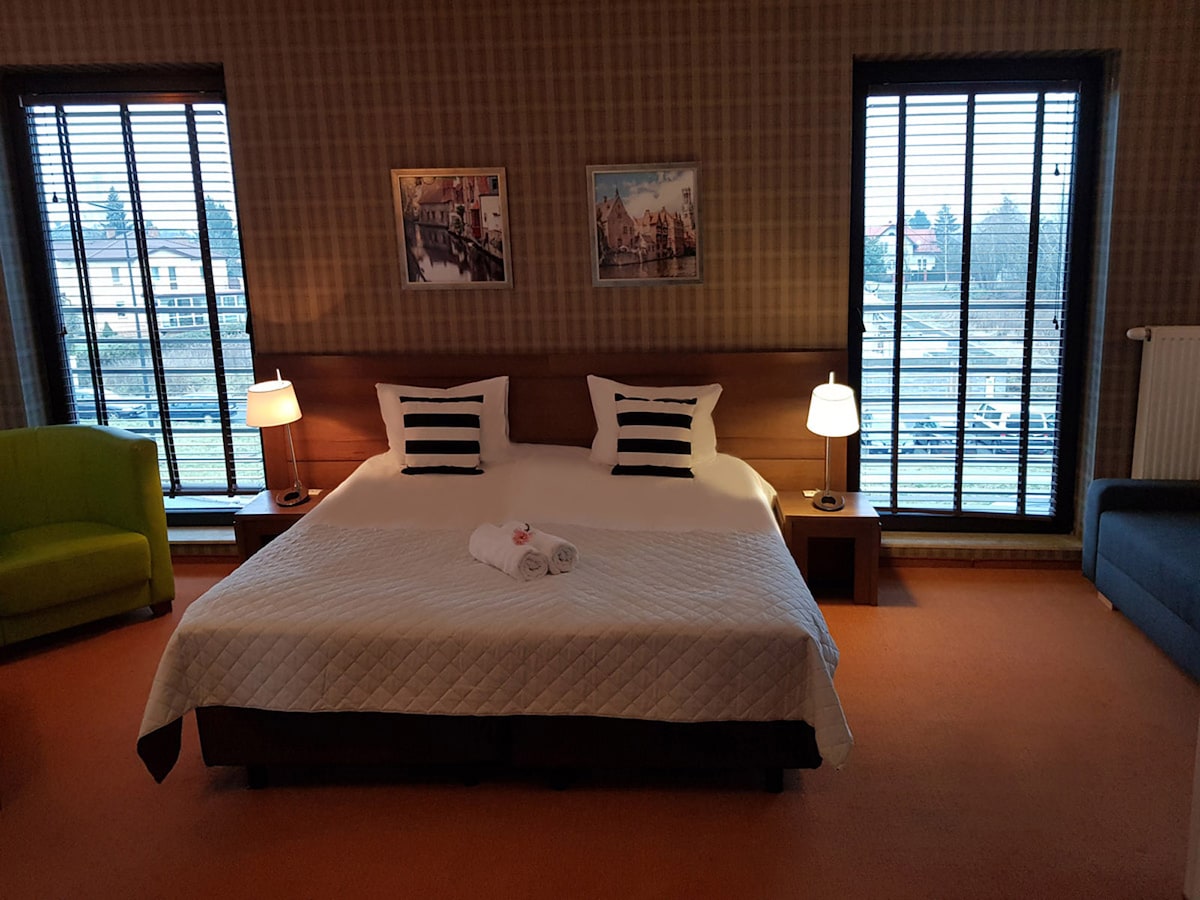 A spacious room features a large bed with crisp linens and decorative pillows. Two bedside lamps provide soft lighting, while large windows with blinds allow natural light to enter. A comfortable armchair and a cozy sofa complete the inviting seating area.