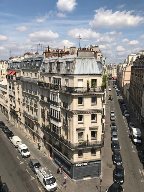 An urban street scene displays a timeless Parisian building with a distinctive, sloped grey roof. Balconies adorned with greenery overlook the street, where parked vehicles line the curb. The sky is filled with fluffy clouds, casting soft shadows across the neighborhood.