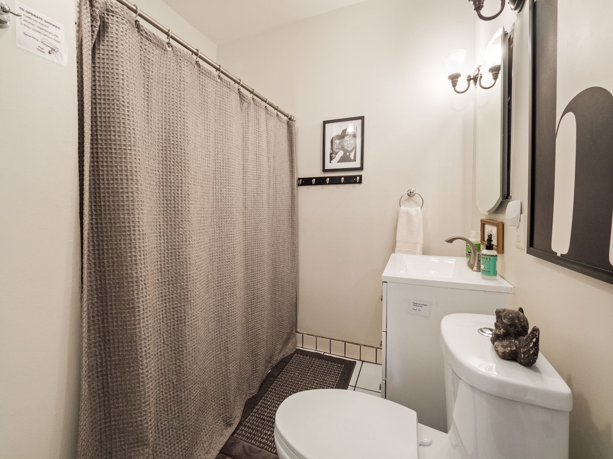 A clean bathroom is presented with a white toilet and a modern sink. A shower area is enclosed by a textured grey curtain. A wall-mounted mirror reflects soft lighting from wall sconces, while a small decorative item sits discreetly on the sink.