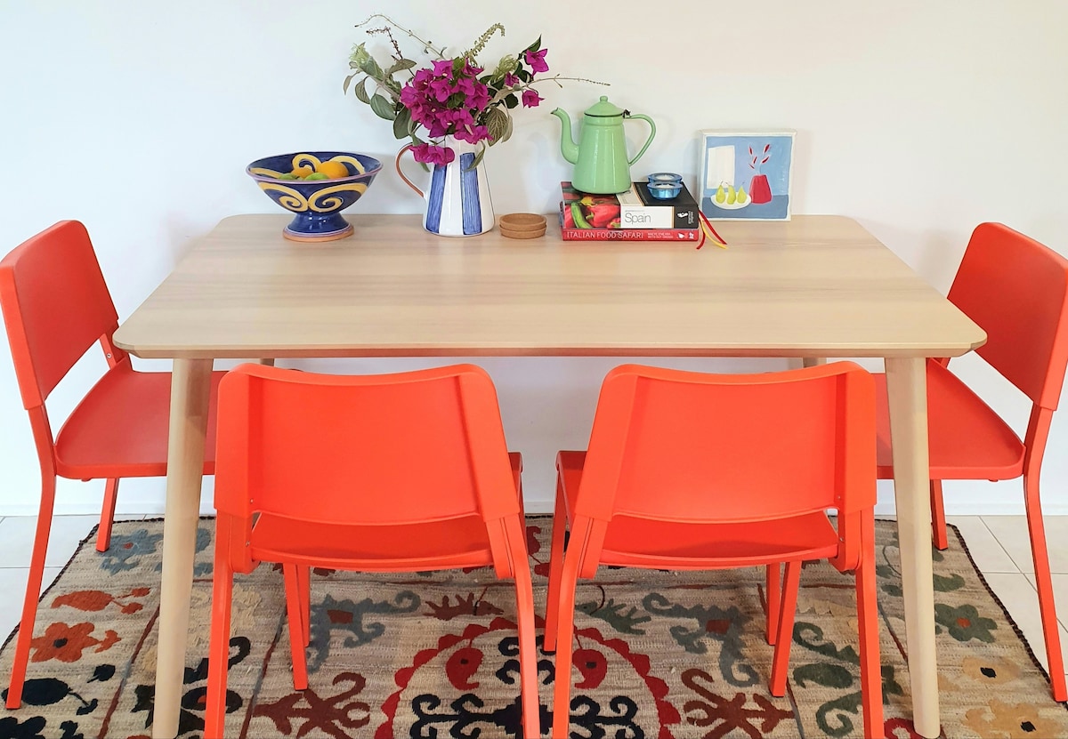 A modern dining area features a light wooden table surrounded by four vibrant orange chairs. A decorative bowl and a small plant adorn the table, alongside a green kettle and a collection of books. The space is anchored by a colorful area rug with intricate patterns.