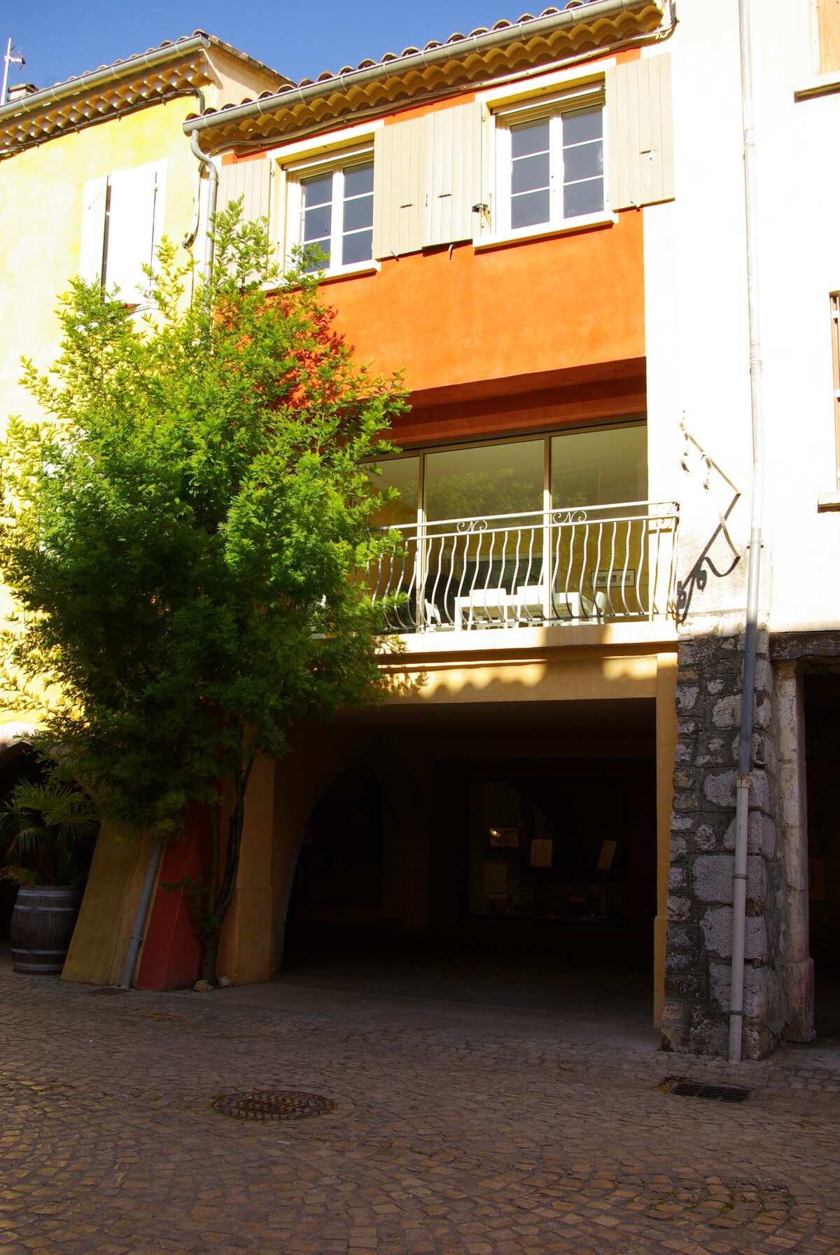 The exterior of a village house is captured, featuring a vibrant orange wall complemented by green shutters. A tree adds greenery near the entrance. The balcony offers a view from the second floor, with large glass doors that provide abundant natural light. Cobblestone streets are visible.