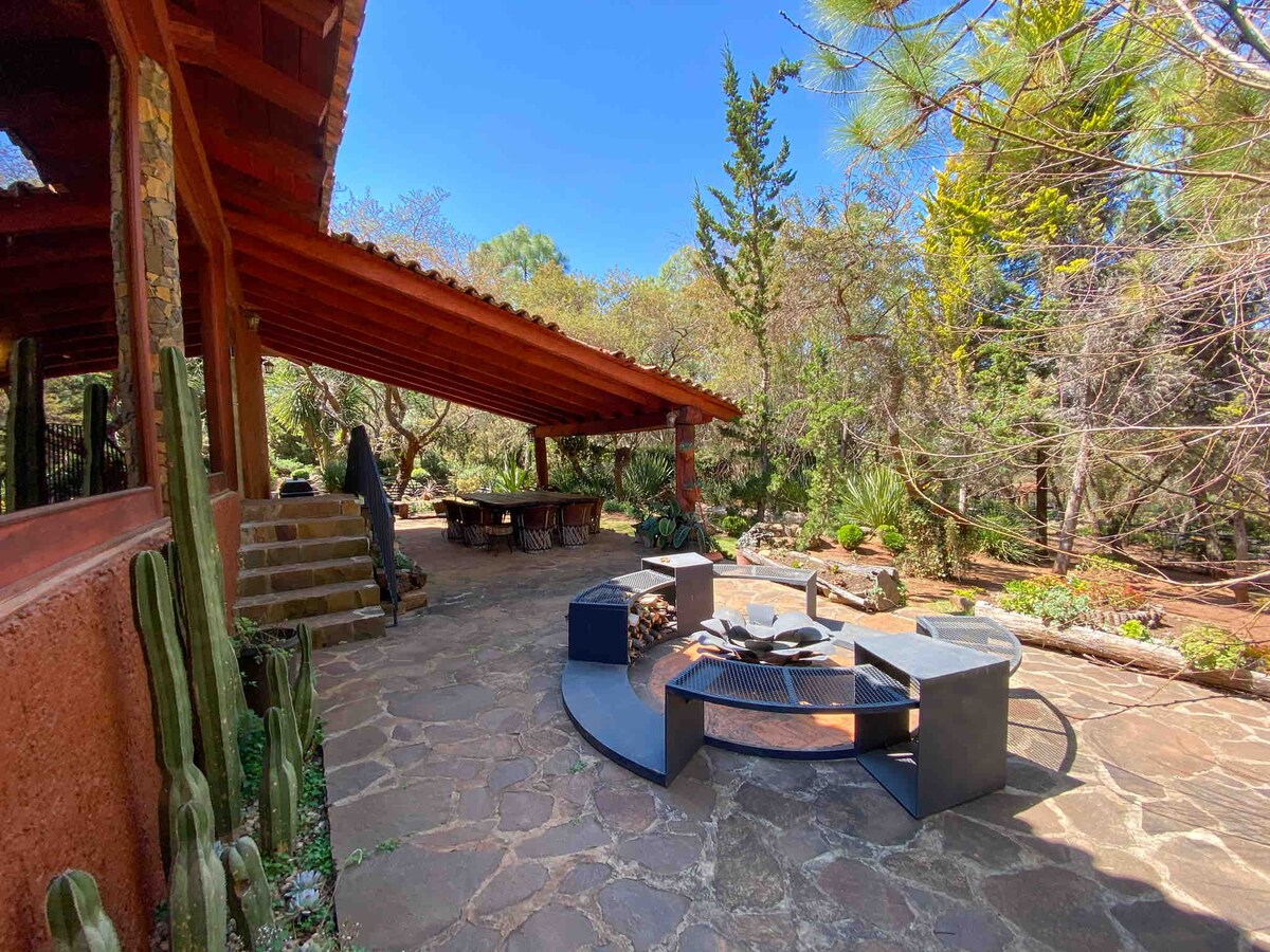 An inviting outdoor area is presented with a spacious stone patio and wooden roof providing shade. A circular fire pit with seating is centrally located. Surrounding greenery, including trees and shrubs, enhances the tranquil atmosphere, creating a perfect spot for relaxation and gatherings.
