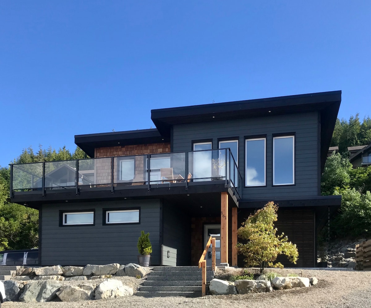 The exterior of a modern, custom-built vacation home is presented, showcasing a dark wooden facade with large glass windows and a spacious deck. A pathway leads to a wooden entrance, bordered by decorative rocks and greenery, under a clear blue sky.