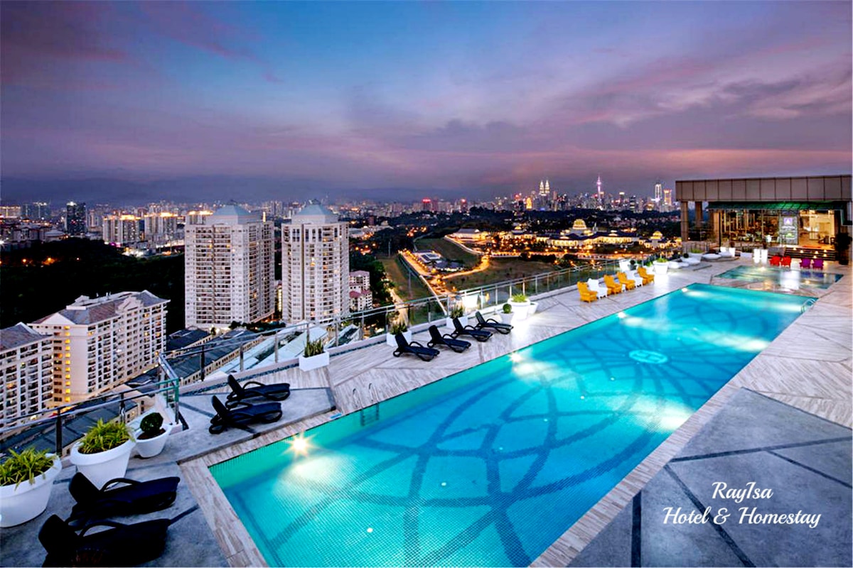 The rooftop pool is surrounded by lounge chairs, providing a view of the city skyline at dusk. The pool's water reflects the colorful evening sky, and the surrounding area features potted plants. A bar area is visible in the background.