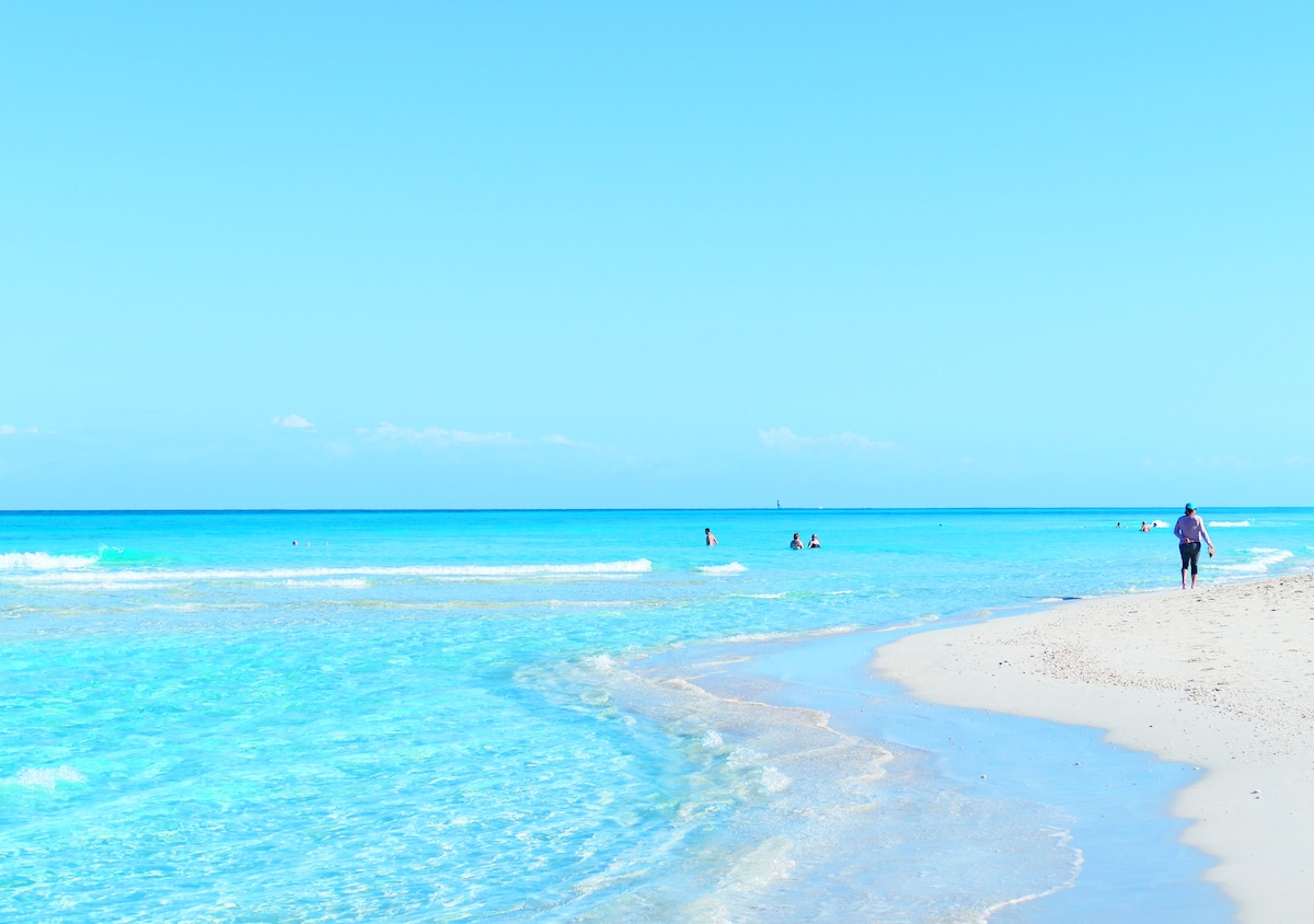 A stretch of sandy beach is depicted alongside tranquil turquoise waters. Gentle waves lap at the shore, while a few people wade into the water. The clear blue sky enhances the peaceful coastal atmosphere.