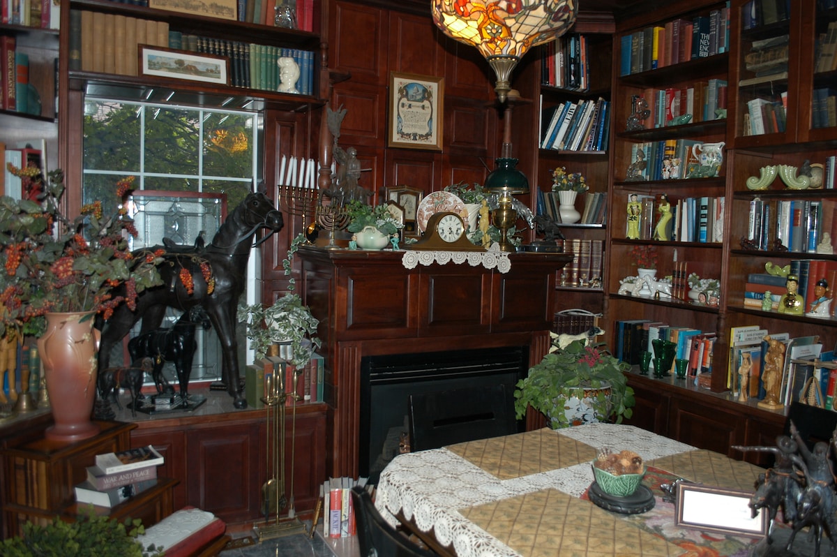 A richly appointed library features dark wooden bookshelves filled with various books and decorative items. A large, stained glass lamp provides ambient light. A cozy seating area is indicated by patterned upholstery, and a fireplace adds warmth to the inviting space.