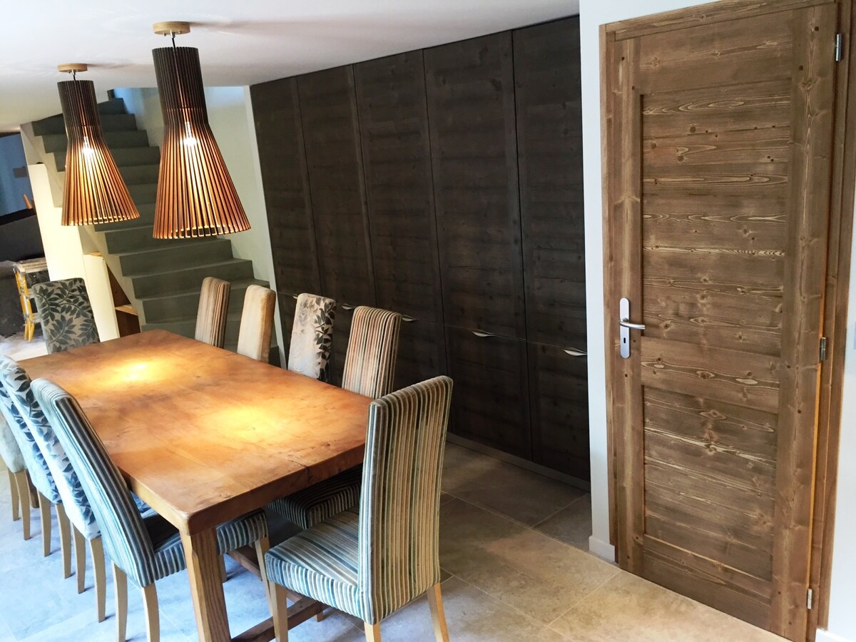 A spacious dining area features a large wooden table surrounded by upholstered chairs, showcasing a mix of striped and patterned fabrics. Two pendant lights hang above the table, providing warm illumination. A modern sliding door is visible, with smooth wooden texture, leading to an adjoining space.
