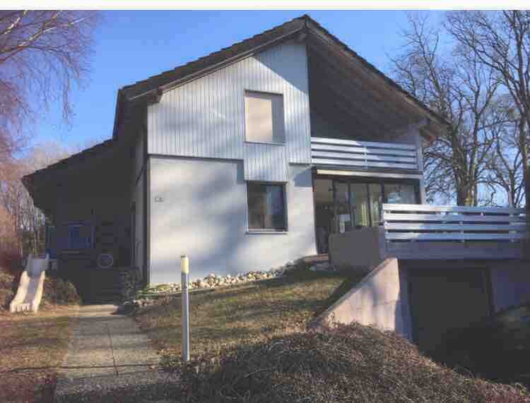 A modern two-story house is presented, featuring a light-colored exterior with wooden accents. Large windows allow natural light to enter, while a small balcony extends from the upper level. The surrounding area includes minimal landscaping with gravel pathways leading to the entrance.