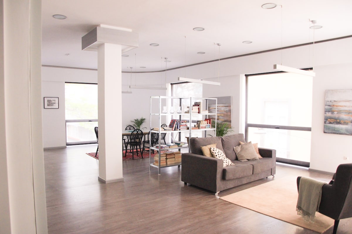 A spacious living area showcases modern design elements, complemented by large windows allowing ample natural light. A cozy gray sofa with decorative pillows invites relaxation. A stylish bookcase divides the room, with a dining table nearby, providing a functional yet open layout.