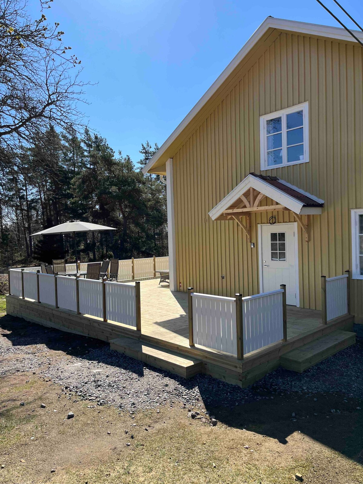 A charming yellow house is framed by a wooden deck featuring a white railing. Sunlight brightens the area, and a shaded outdoor space is provided by an umbrella. Trees stretch in the background, enhancing the natural surroundings.
