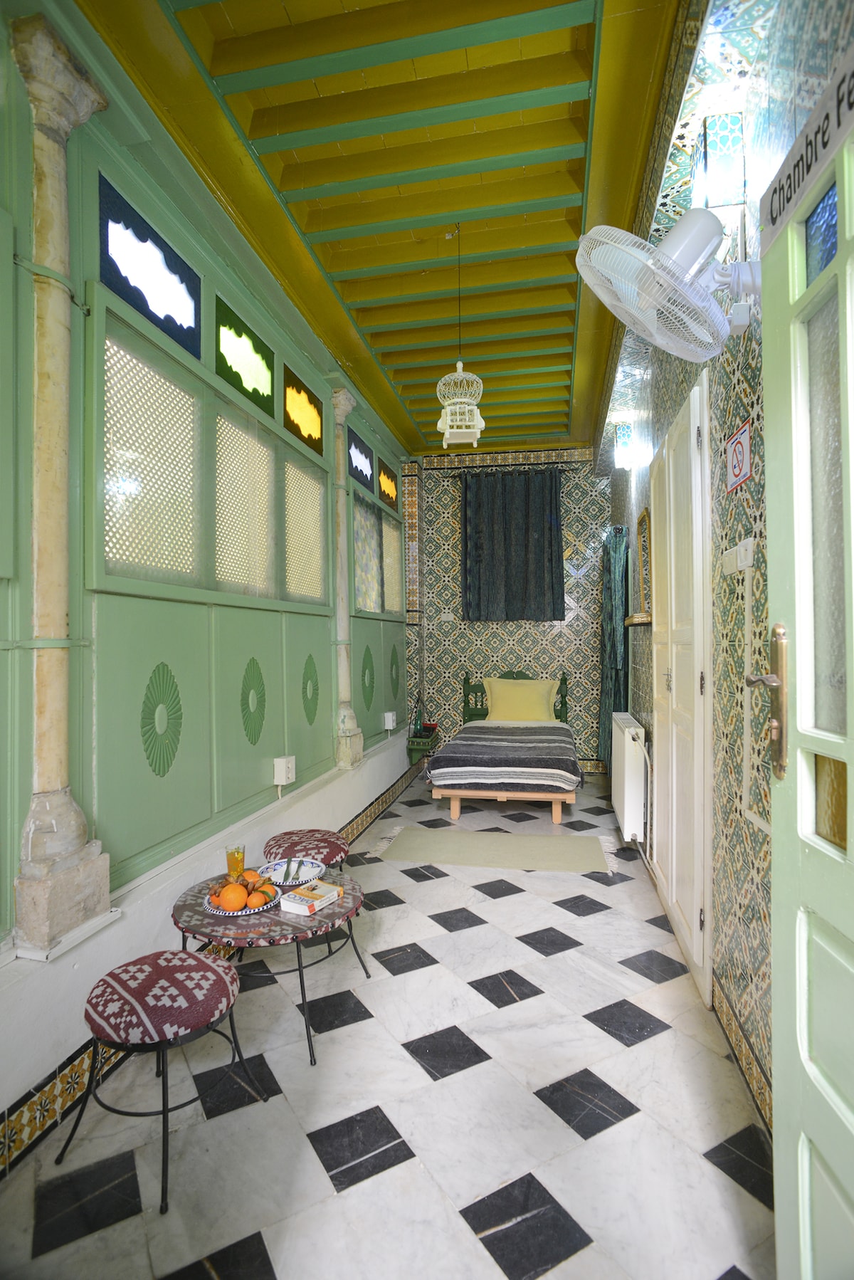 A hallway features patterned tile flooring, with a cozy sleeping area in the background. The spacious bed is neatly made, surrounded by decorative mosaic tiles. A small table with assorted fruit and two patterned chairs are positioned nearby, under colorful glass window panels.