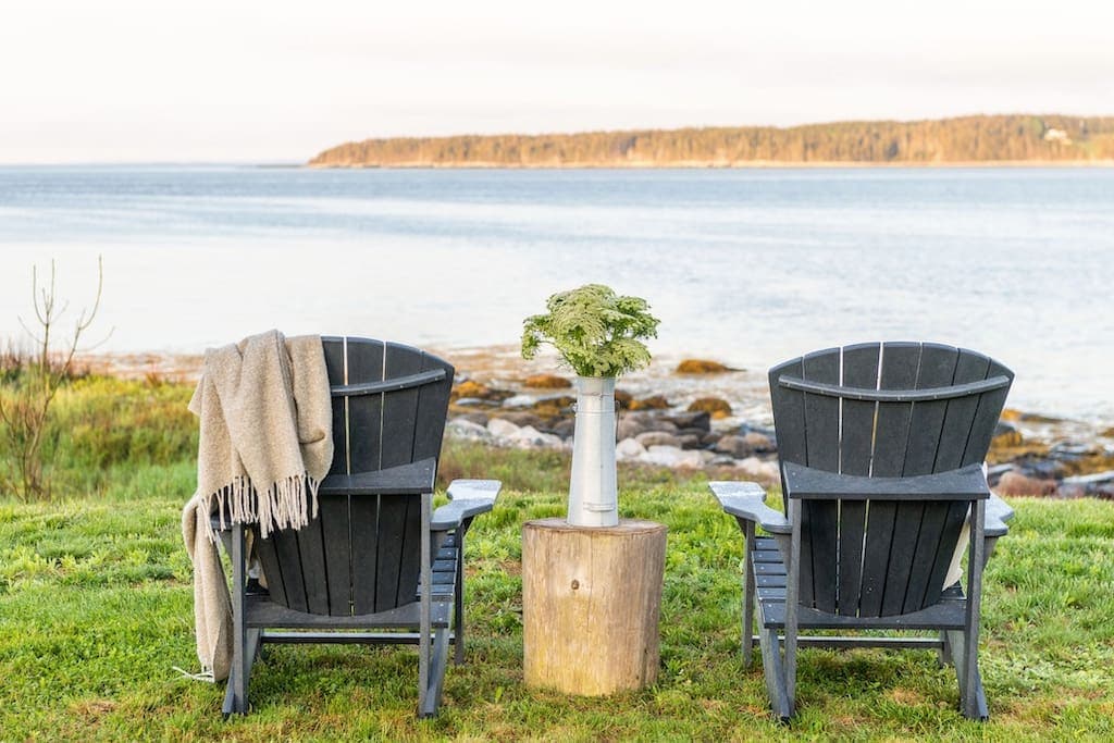 Two dark wooden adirondack chairs are positioned by the water, facing the ocean. A small wooden table sits between them, holding a vase with flowers. A soft blanket drapes over one chair, and a peaceful coastal view displays gently rolling waves and distant tree-lined hills.