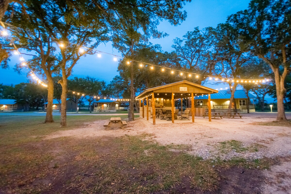 A spacious outdoor area is illuminated by string lights, creating a soft glow around a covered pavilion. Nearby picnic tables are arranged on gravel, surrounded by trees. The backdrop features cottage-style buildings, enhancing the serene setting at twilight.
