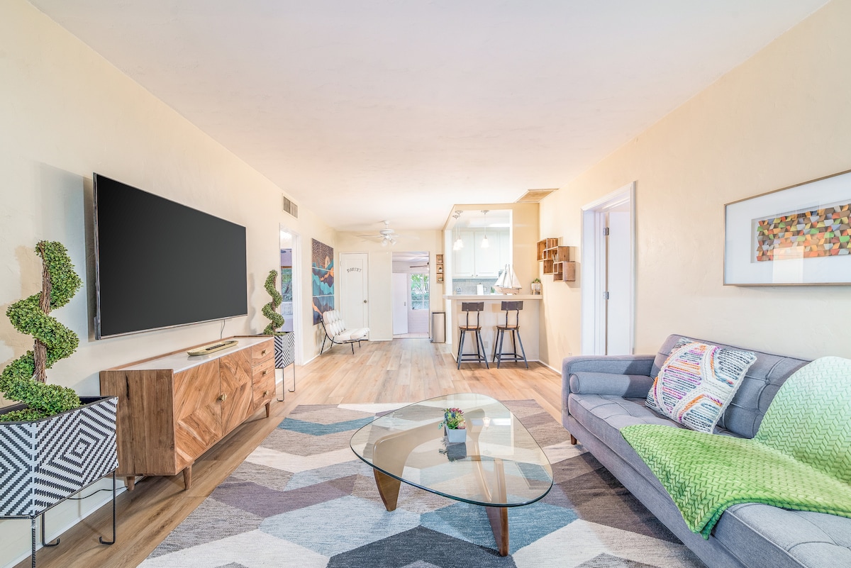 A spacious living area features a modern glass coffee table at the center, surrounded by a patterned area rug. A comfortable sofa sits adjacent to a wooden sideboard, which supports a large flat-screen television. Natural light filters in, suggesting an inviting atmosphere.