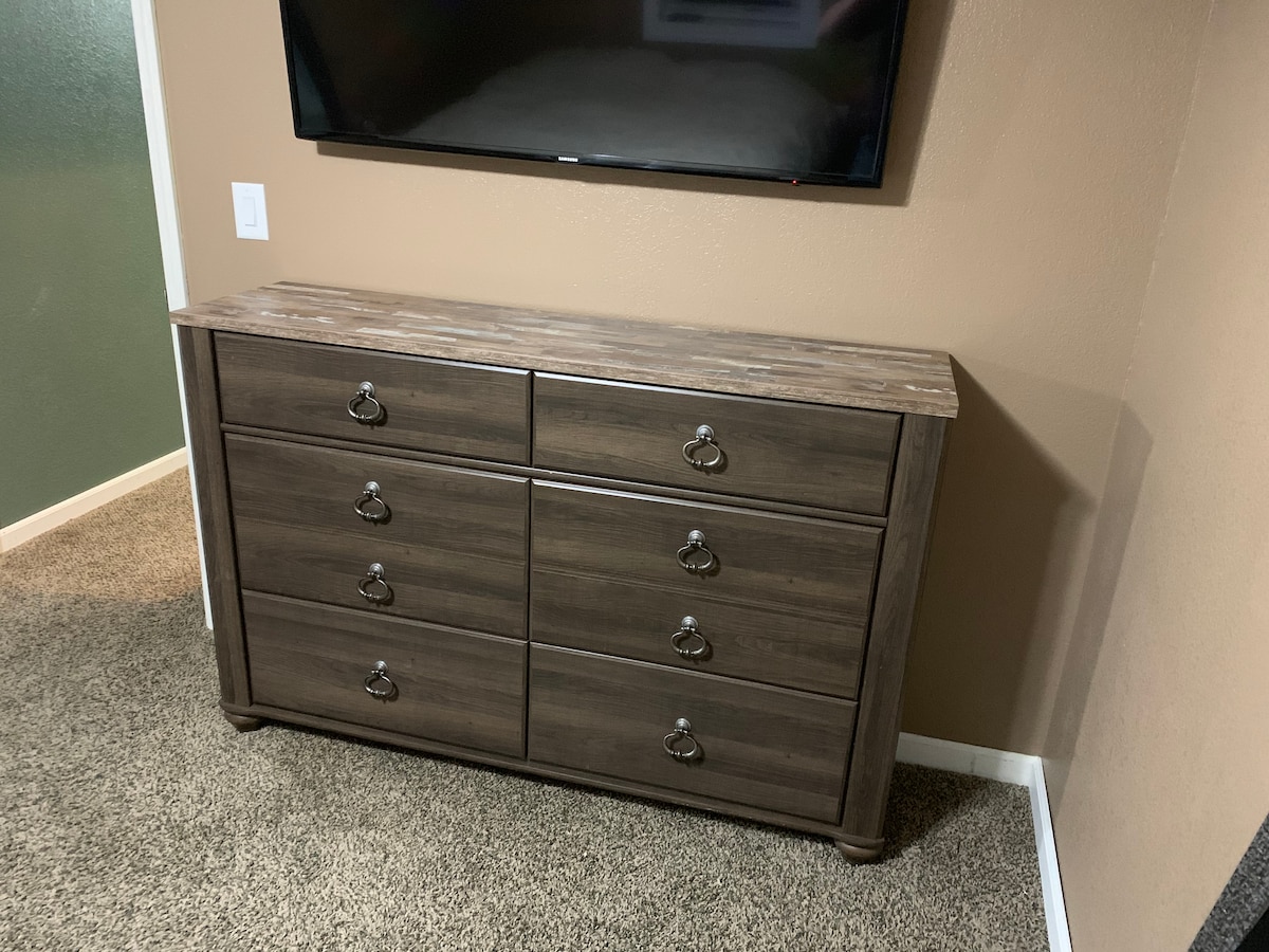 A wooden dresser with a sleek design features six drawers adorned with metal handles. A flat-screen television is mounted above the dresser, positioned against a neutral-colored wall, contributing to the room's functional layout.