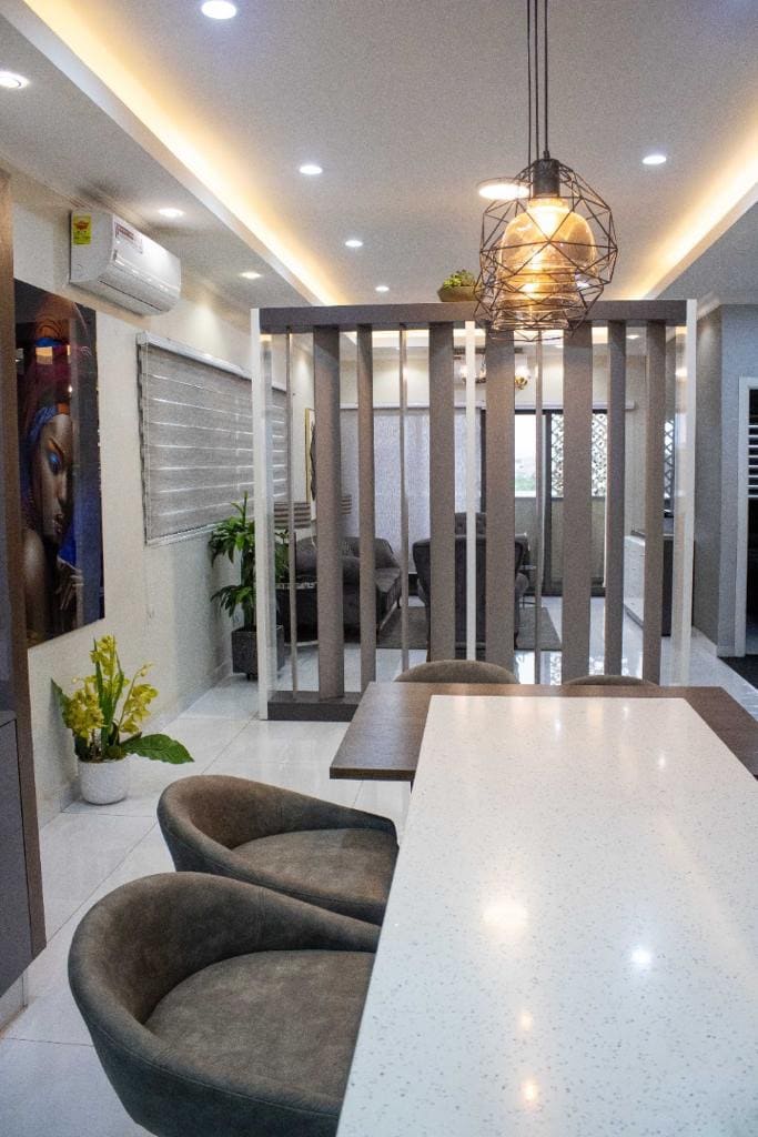 An open layout is seen featuring a kitchen island with a speckled countertop. Plush, rounded chairs are positioned around the dining area. Dividing the space, wooden slats create a cozy separation leading to a lounge area visible in the background, illuminated by soft ceiling lights.