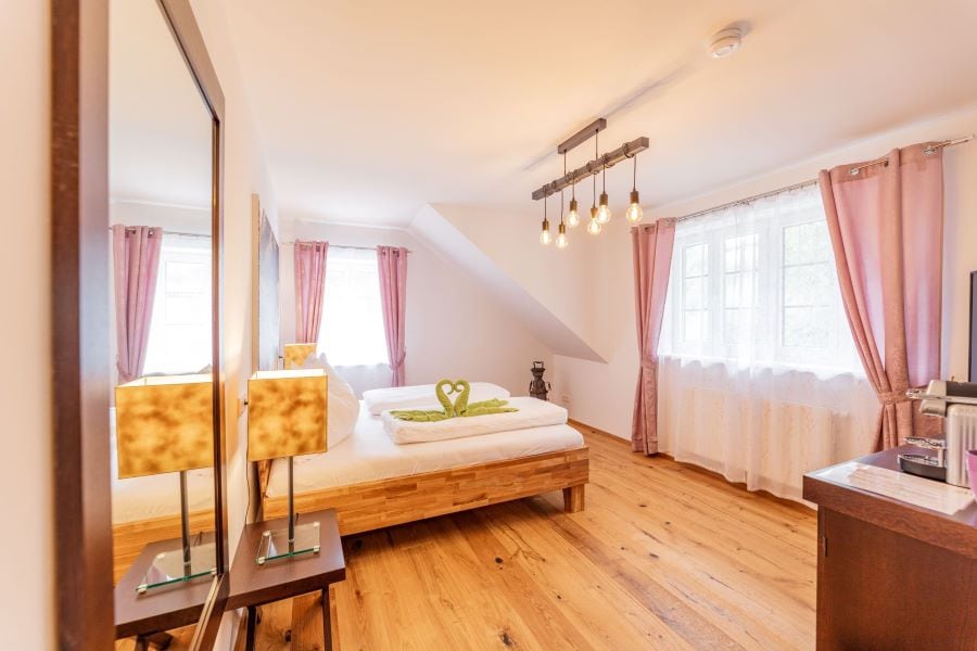 A bright and airy room features a spacious bed with a green accent thrown over the top. Warm wooden flooring complements the soft glow from hanging light fixtures. Two windows, dressed in light curtains, allow natural light to fill the space, enhancing the inviting ambiance.