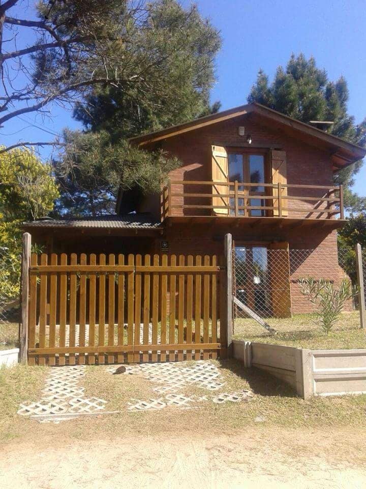 A two-story wooden cabin is viewed from the front, displaying a fenced yard and a gravel pathway leading to the entrance. The upper level features a balcony enclosed by wooden railings, while large windows allow natural light to enter the space.