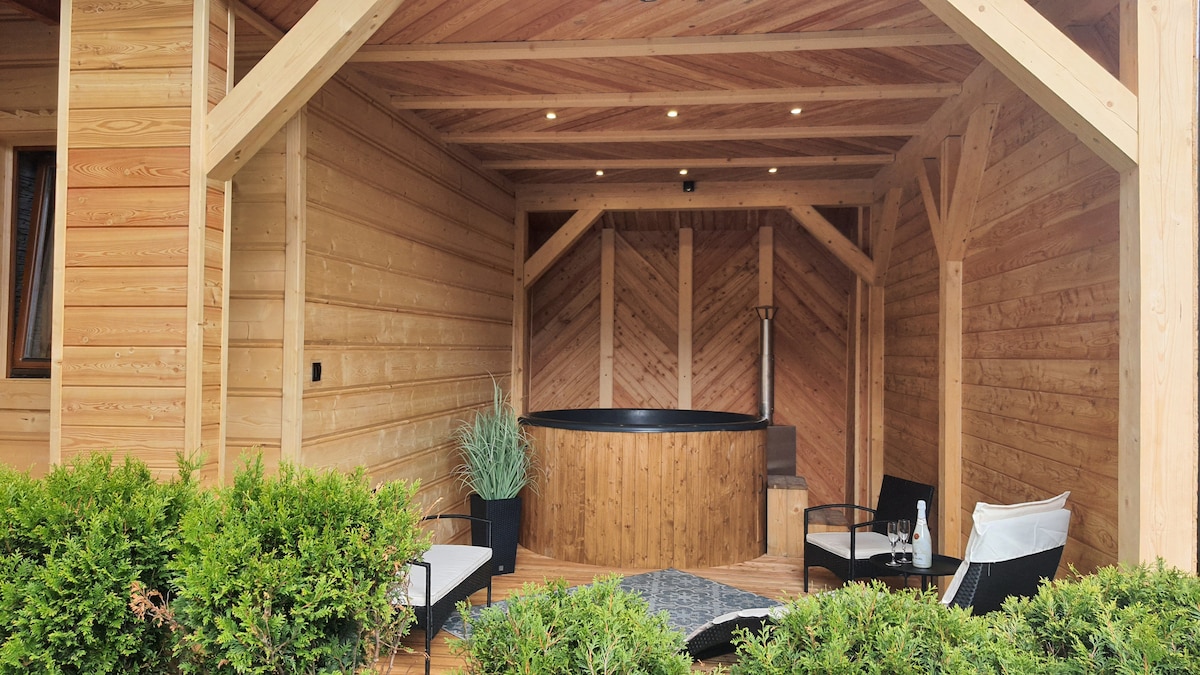 An outdoor area features a round wooden jacuzzi surrounded by wooden beams and a deck. Comfortable black seating is present beside the jacuzzi. Lush greenery borders the space, creating a serene atmosphere for relaxation.