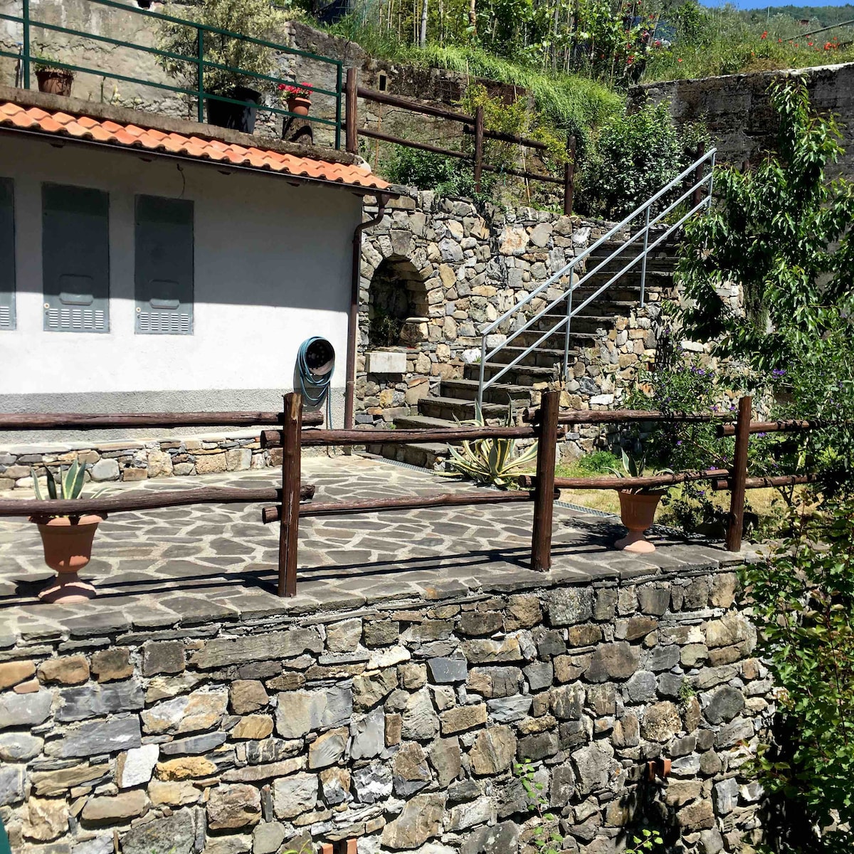 An outdoor area is shown, featuring a stone terrace surrounded by greenery. Wooden railings encircle the space, with potted plants positioned thoughtfully. A set of stairs leads to an upper level, and a rustic stone wall complements the tranquil setting.