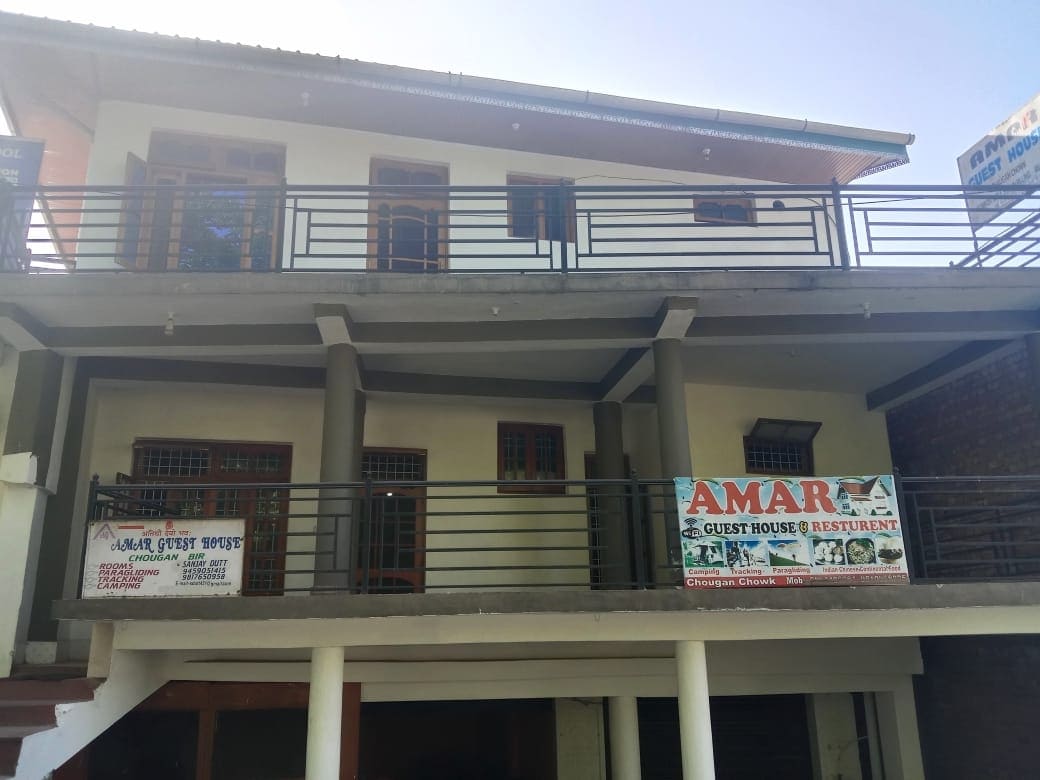 A two-story building is featured with a covered balcony and large windows. A sign indicates the presence of Amar Guest House and Restaurant. The restaurant's name is prominently displayed, inviting guests to enjoy the amenities offered.