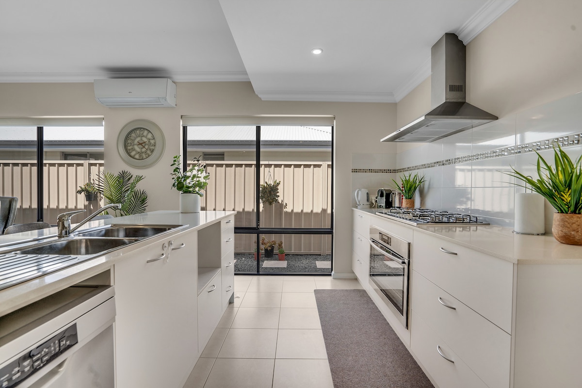 A bright, modern kitchen features a spacious island with seating, stainless steel appliances, and ample counter space. Large windows allow natural light to fill the room, showcasing a view of the outdoor area. Potted plants add a touch of greenery to the neutral decor.
