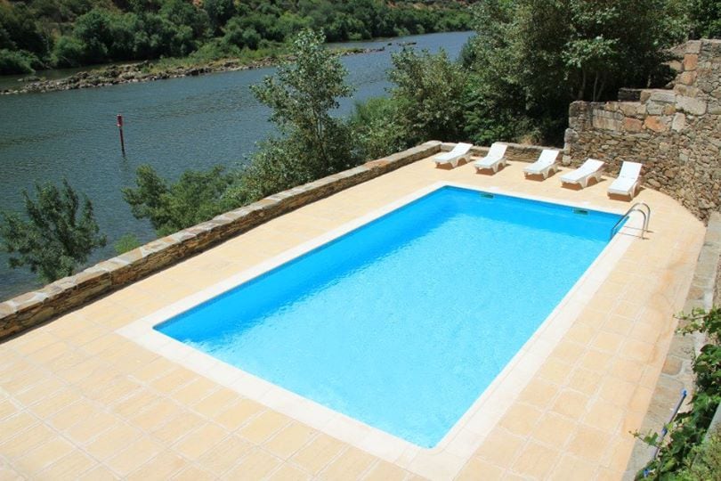 A rectangular swimming pool features clear blue water surrounded by a sunbathing area. Four lounge chairs are positioned along one side, with lush greenery and the Douro River visible in the background, enhancing the serene outdoor setting.