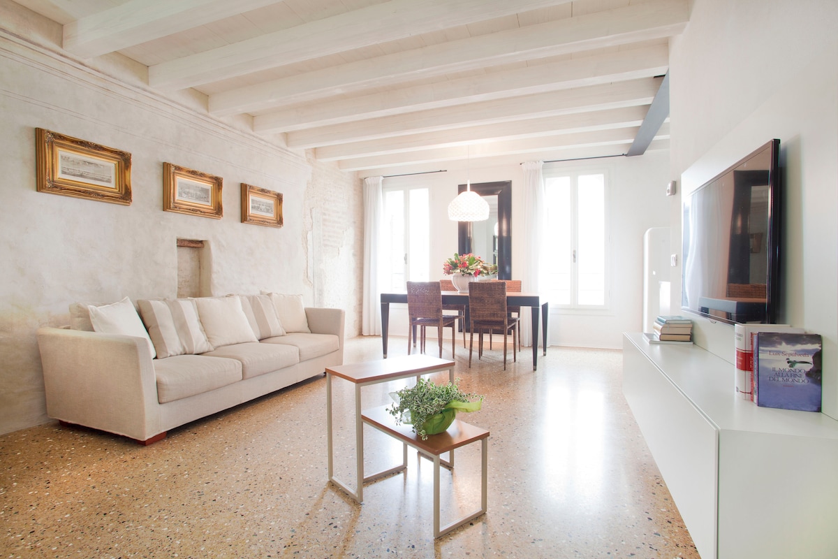 A spacious living area features a light-colored sofa and a modern coffee table. Natural light fills the space through large windows, highlighting the exposed wooden beams and textured walls. A dining table with four chairs is set for meals, with a decorative vase in the center.