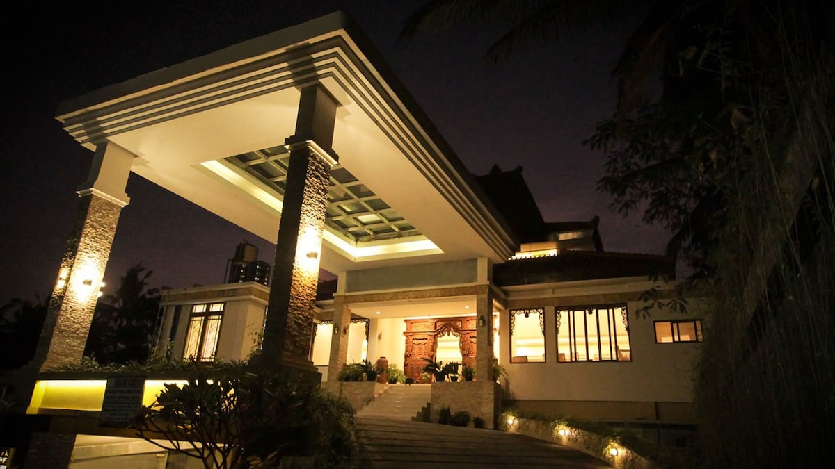 The villa is showcased at night, featuring a grand entrance with a modern overhang and stone accents. Soft lighting highlights the pathway leading to the entrance, while large windows provide glimpses of the interior. The architecture reflects a blend of contemporary and traditional Javanese design.