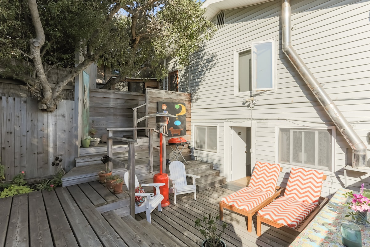 An outdoor deck area is presented, featuring two lounge chairs with orange and white chevron patterns. A wooden staircase leads up to the entrance. Decorative potted plants and a painting are visible on the wooden fence and wall, enclosed by greenery providing a sense of privacy.