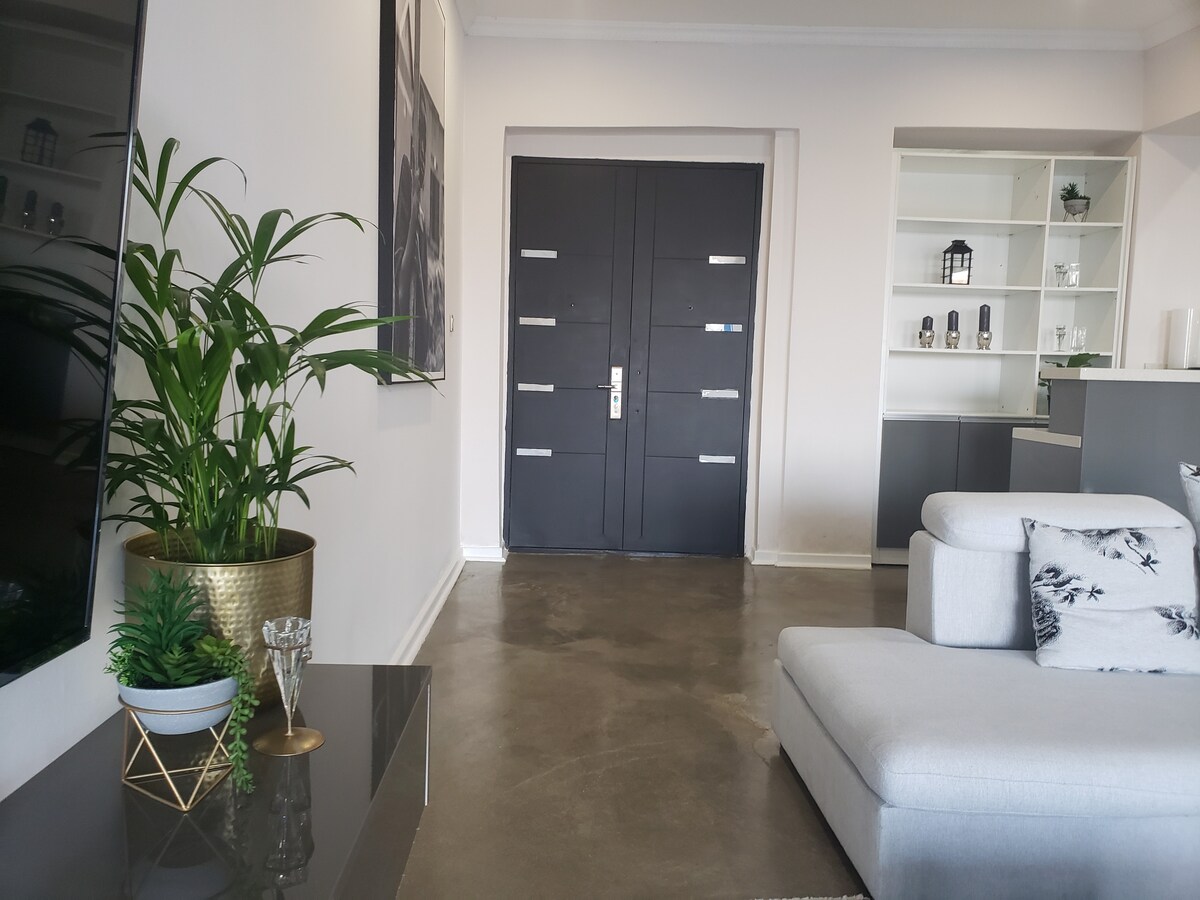 An entrance area showcases a spacious, modern gray sofa positioned next to a glass coffee table. A green potted plant adds a touch of nature, while large double doors with sleek metal handles serve as a focal point. Minimalist shelving displays decorative items.