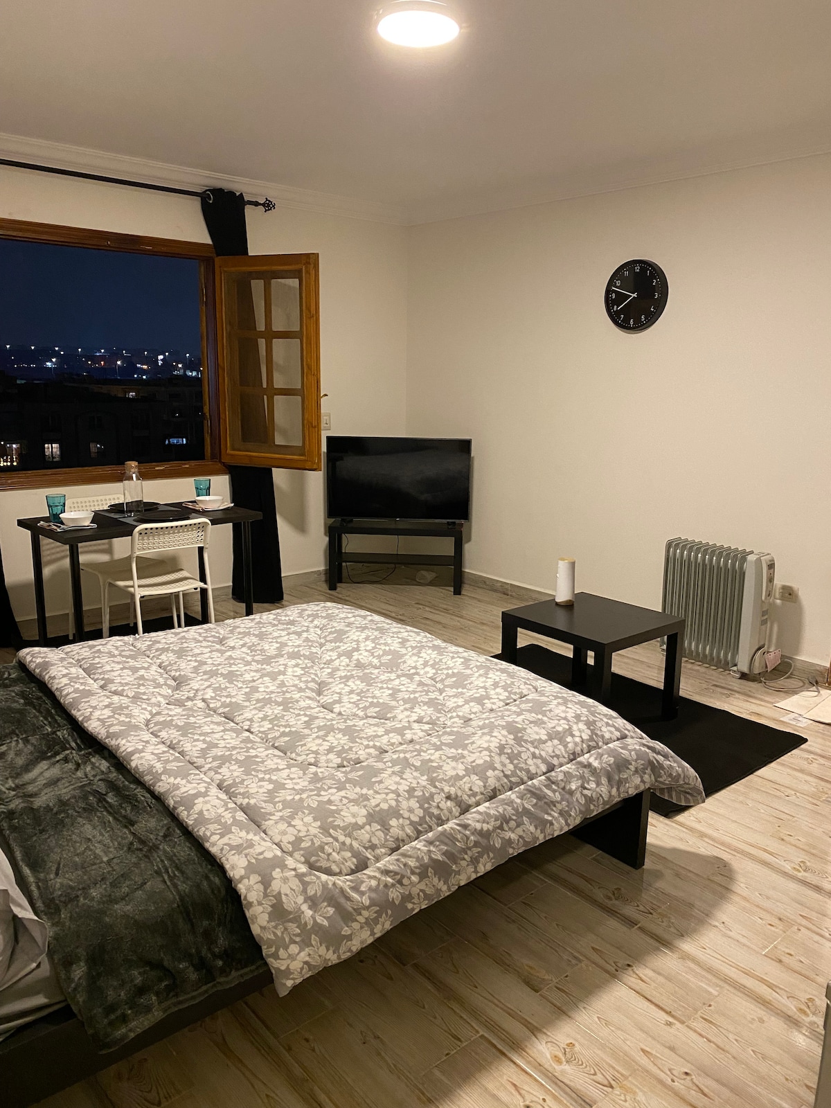 A spacious room is seen featuring a large bed with a floral-patterned blanket, a small dining table with two chairs, and a television on a stand. A window allows for city views, while a clock adorns the wall. A heater provides additional comfort.