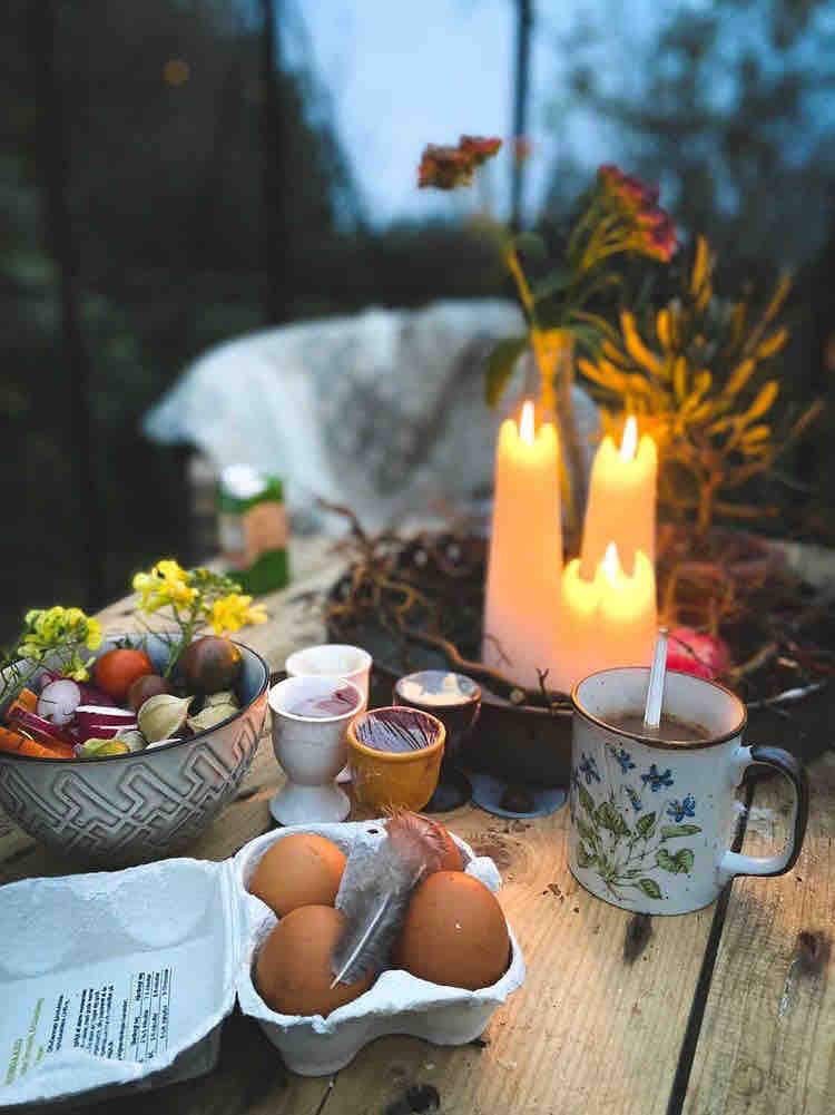 A wooden table is set with a variety of fresh produce, including eggs and sprigs of flowers. Two lit candles provide soft illumination alongside a cup of coffee. Small decorative items, such as colorful eggs, enhance the tranquil scene.