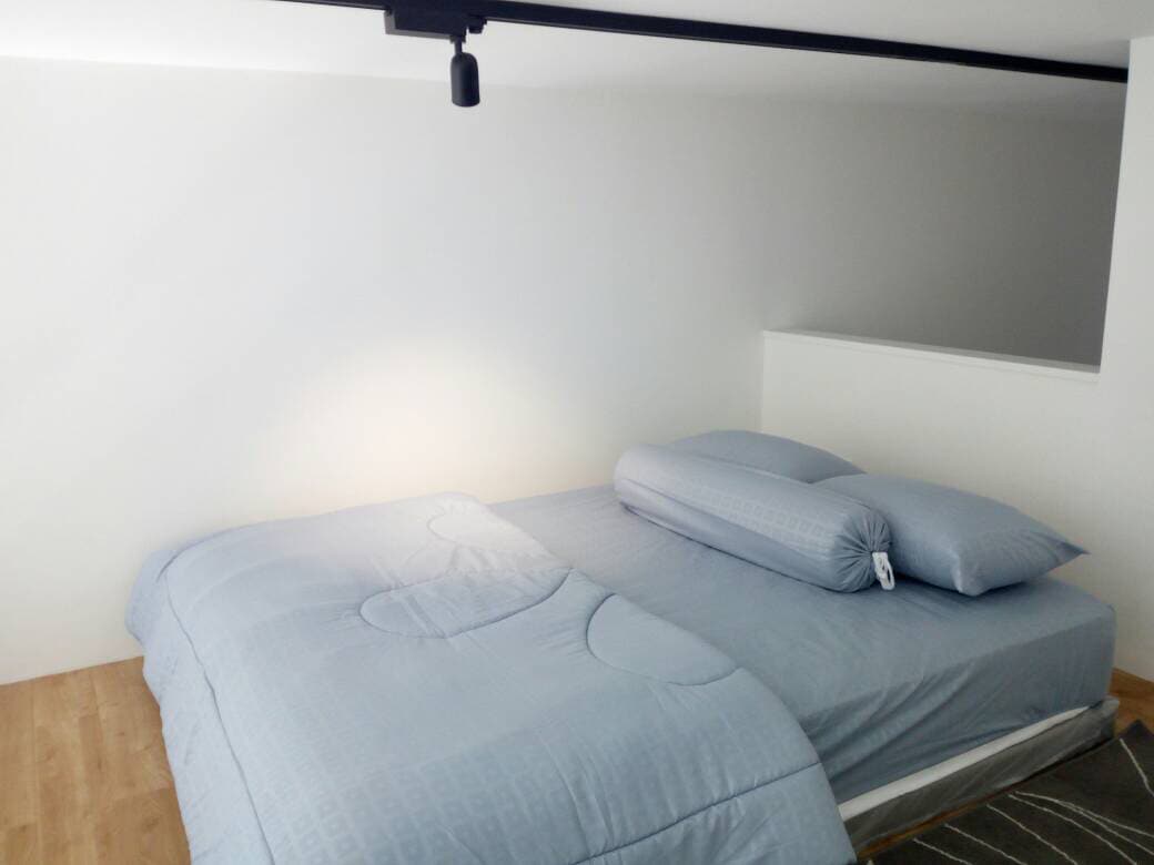 A loft-style sleeping area features a spacious bed with soft blue bedding and a matching bolster pillow. The walls are painted white, and natural light softly illuminates the space from above, highlighting the warm wooden flooring.