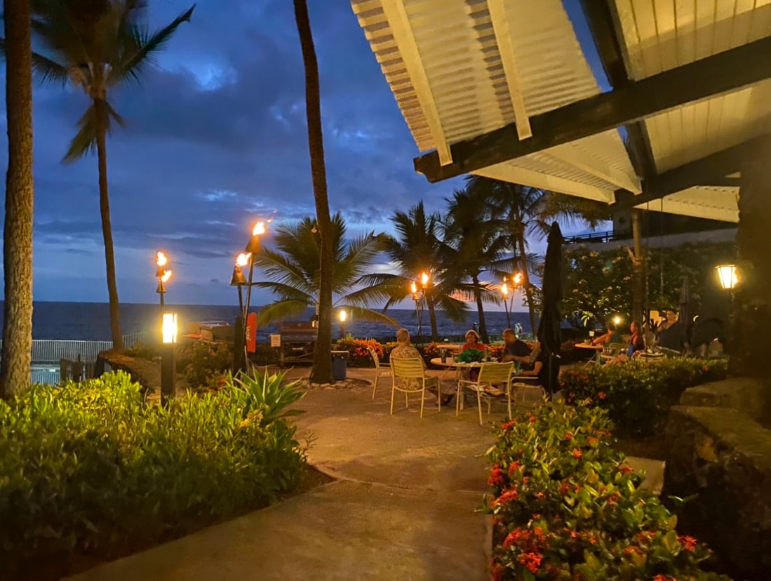 A serene outdoor dining area is illuminated by tiki torches against a backdrop of palm trees. A pathway guides guests through lush tropical gardens towards seating arrangements, with the ocean visible in the distance under a twilight sky.
