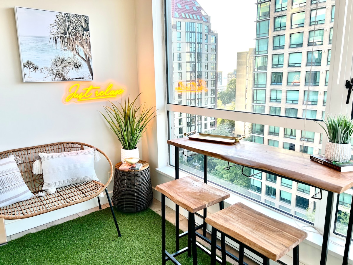 A bright den/solarium features large windows offering views of downtown buildings. A rattan chair with soft cushions sits alongside a wooden table and two stools. A small potted plant and a neon sign saying 'Just Relax' add character to the space.