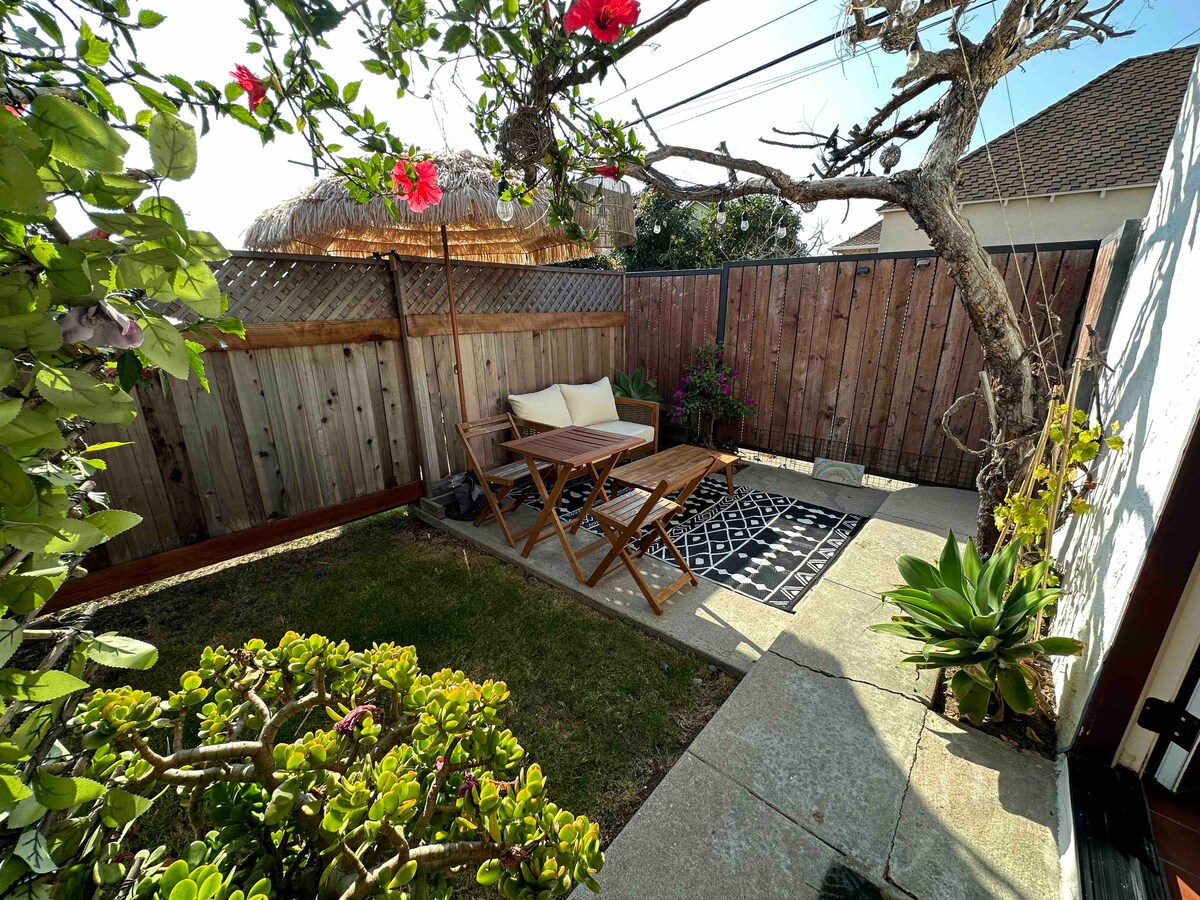 A serene outdoor space is highlighted with a decorative rug on a stone patio. A wooden table and chairs are positioned for relaxation, surrounded by well-maintained greenery and vibrant flowers. A thatched umbrella provides shade, creating an inviting atmosphere.