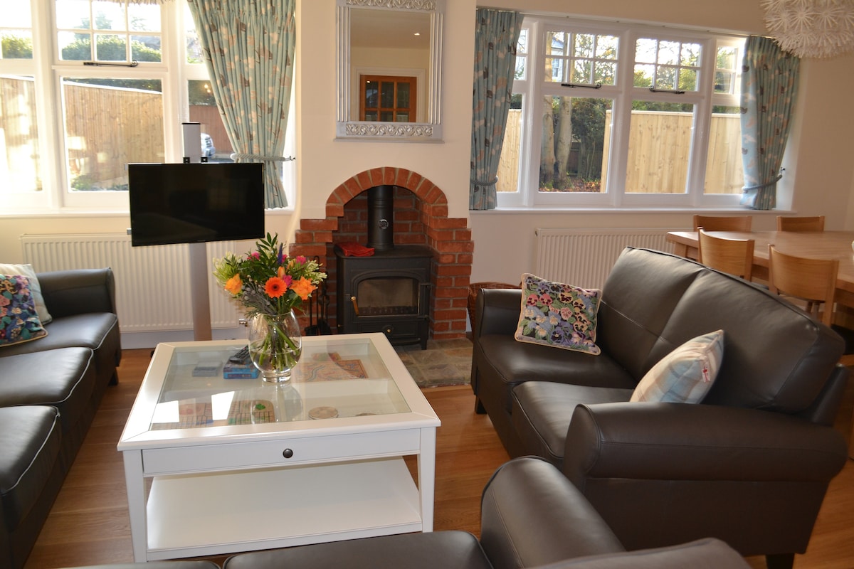 A comfortable lounge area features a white coffee table with a floral arrangement and a cozy wood burner in a brick hearth. Two dark leather sofas are arranged around the table, while a dining table with wooden chairs is visible in the background near large windows.