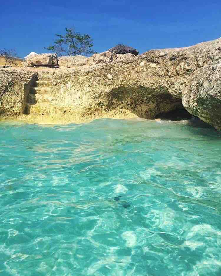 Clear turquoise water gently laps against rocky shorelines, revealing natural textures and colors. Sunlight dances on the water's surface, creating a serene atmosphere. Soft, sandy steps lead into the inviting water, surrounded by jagged rocks and sparse greenery.