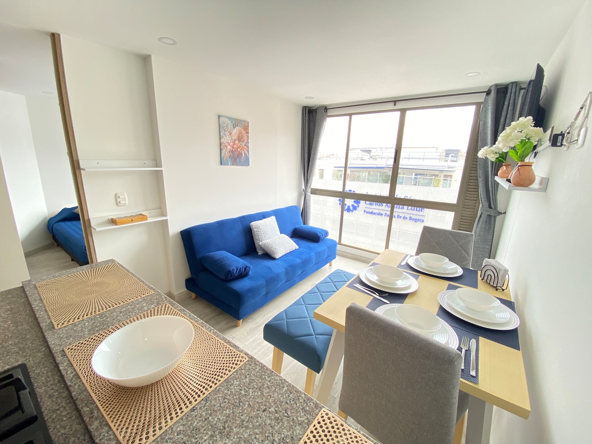 A bright living area features a blue sofa and a dining table set for four, with plates and utensils arranged neatly. Large windows provide natural light and views of the outside. The open layout connects to a cozy sleeping area visible in the background.