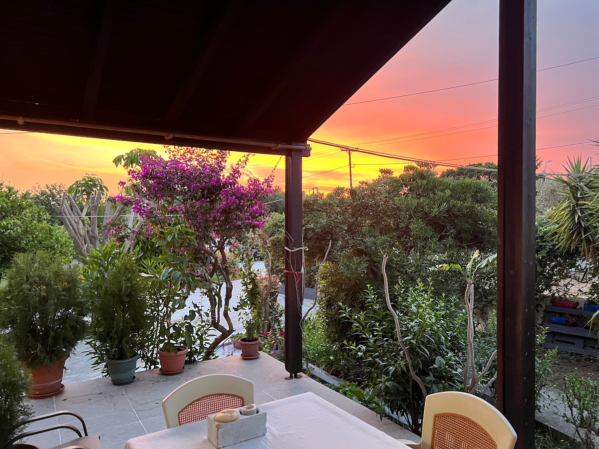 A serene outdoor dining area is visible, framed by wooden beams. Potted plants and vibrant flowers enhance the space. A sunset casts warm hues of orange and pink across the sky, creating a peaceful backdrop for evening meals.