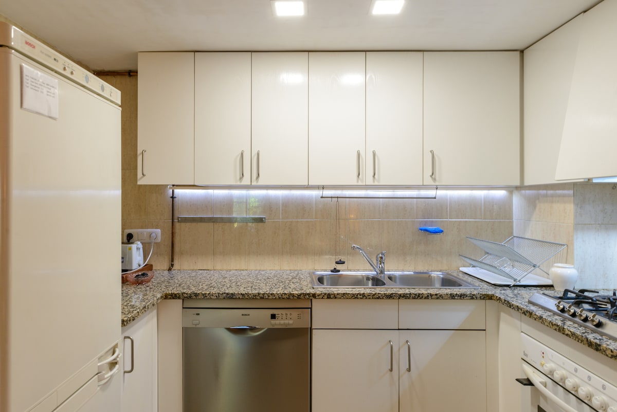 A modern kitchen setup is displayed, featuring sleek white cabinetry and a granite countertop. Stainless steel appliances, including a dishwasher and stove, are neatly integrated. Soft under-cabinet lighting illuminates the workspace, and a dish drying rack is positioned next to the sink.