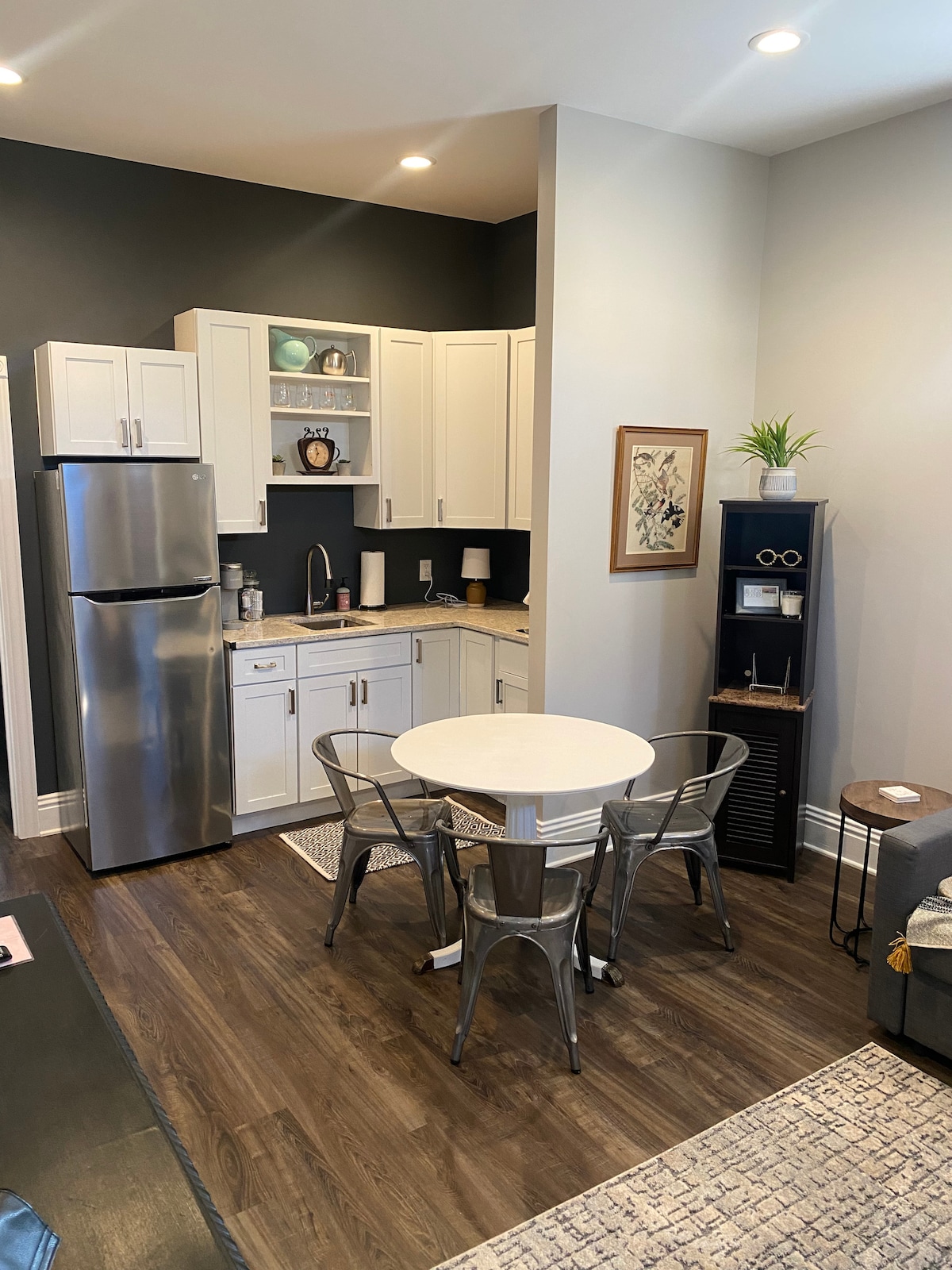 A modern kitchenette is visible, featuring stainless steel appliances, including a refrigerator and an induction cooktop. A round dining table with metal chairs occupies the center space, while decorative items add a touch of style to the cabinetry. Soft lighting enhances the warm tones of the wood floor.