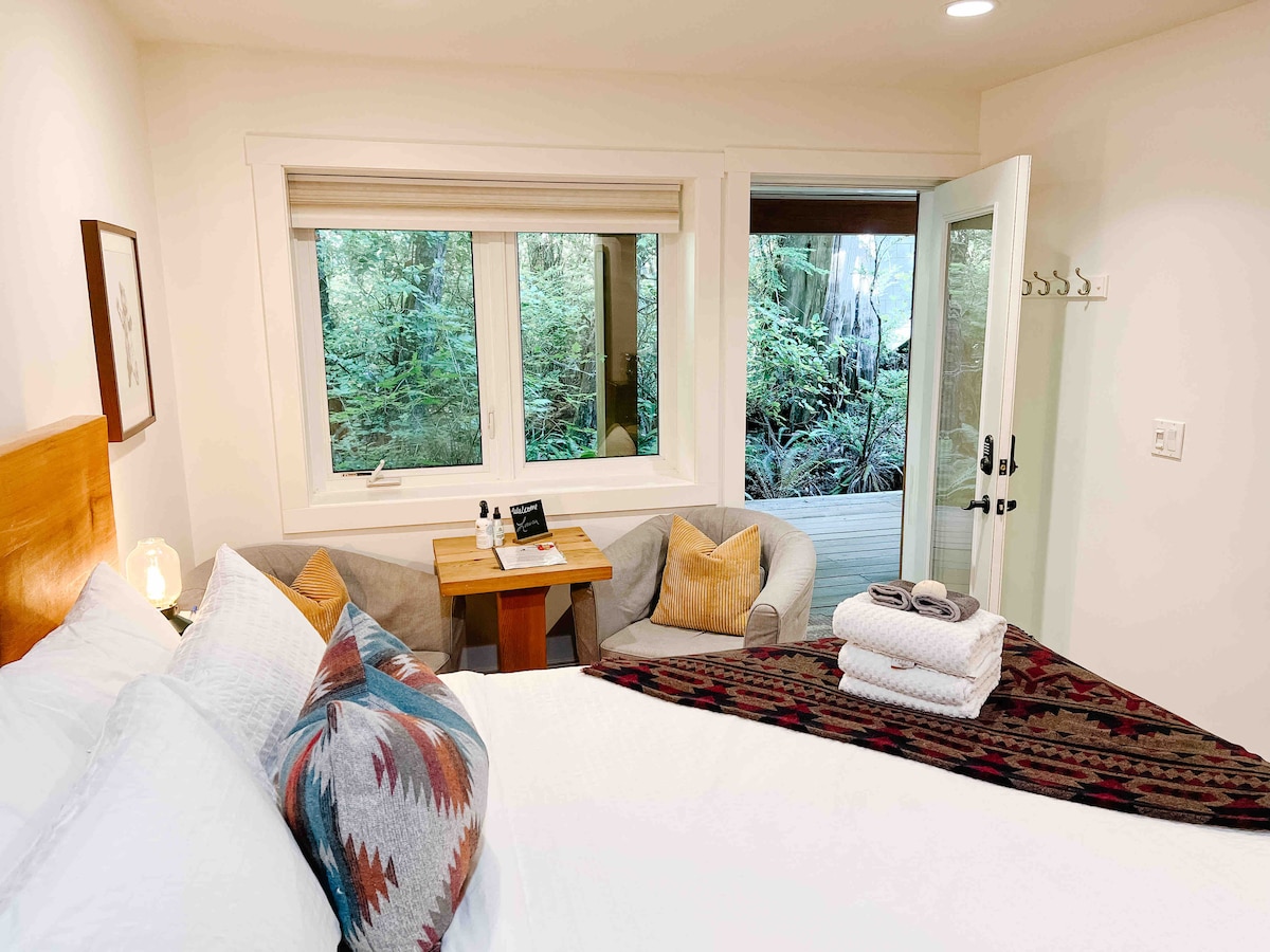 A cozy studio suite features a spacious bed adorned with decorative pillows. Natural light enters through large windows, offering views of the surrounding rainforest. A seating area is visible, and a small table is positioned beside it, creating an inviting space for relaxation.